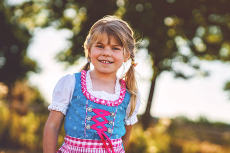 Cute little kid girl in traditional Bavarian costume in wheat field. Preschool girl play at hay bales during summer harvest time in Germany.の写真素材