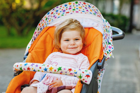 Portrait of little cute smiling toddler girl sitting in stroller or pram and going for a walk. Happy...