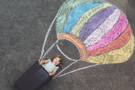 Happy little toddler girl flying in hot air balloon painted with colorful chalks in rainbow colors on ground or asphalt in summer. Cute child having fun. Creative leisure for kids.の写真素材