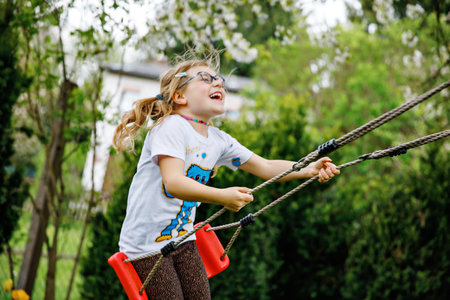 Happy little preschool girl with glasses having fun on swing in domestic garden. Cute healthy child...