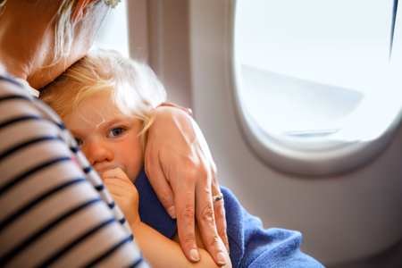 Woman traveling with little child by airplane. Sad tired toddler girl sitting with mum by aircraft window. Motherhood concept. Crying baby.の写真素材