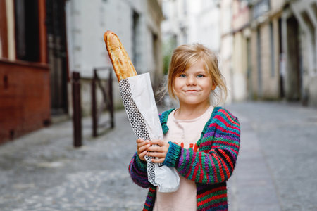 Adorable little preschool girl with fresh French baguette on the street side of the city. Happy small child in Paris, France.の写真素材