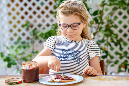 Cute funny preschool girl eats sweet bun for breakfast. Happy child eating bread roll with strawberry, cherry or plum jam. Health food for children and kids with selfmade jellyの写真素材