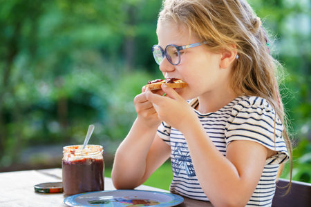 Cute funny preschool girl eats sweet bun for breakfast. Happy child eating bread roll with strawberry, cherry or plum jam. Health food for children and kids with selfmade jellyの写真素材