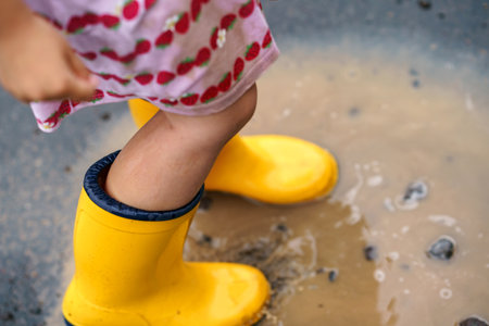 Close-up of little preschool girl wearing yellow rain boots and walking during puddles. Cute child in colorful clothes jumping into puddle, splashing with water, outdoor activityの写真素材