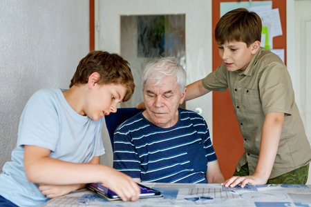 Two Preteen Boys Teaching Grandfather How to Use Internet Safely. Teenage Brothers, School Children with Digital Tablet Playing with Granddad, Senior Man at Homeの写真素材