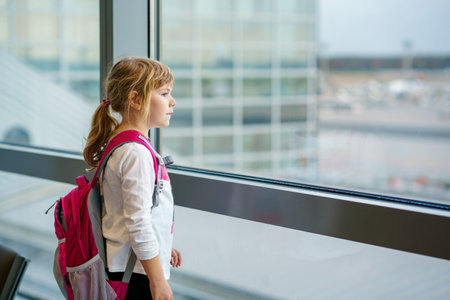 Little Girl at the Airport Waiting for Boarding at the Big Window. Cute Kid Stands at the Window against the Backdrop of Airplanes. Looking Forward to Leaving for a Family Summer Vacation.の写真素材