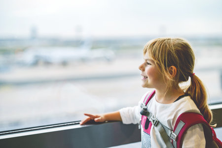 Little Girl at the Airport Waiting for Boarding at the Big Window. Cute Kid Stands at the Window against the Backdrop of Airplanes. Looking Forward to Leaving for a Family Summer Vacation.の写真素材
