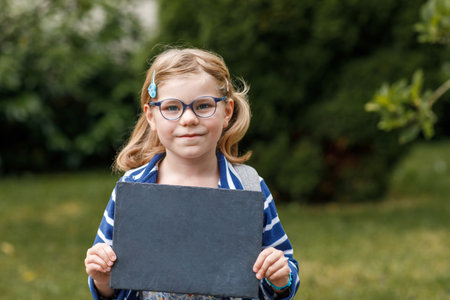 Happy little kid girl standing holding chalkboard in hands. Empty board for copy space. Healthy preschool child wearing eyeglasses outdoors, in green park.の写真素材