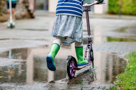 Girl on a scooter in rainy weather. Child in gum rainboots riding through puddles after summer rain. Fun for children.の写真素材