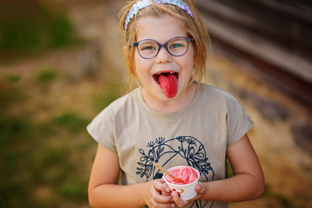 Little adorable preschool girl with eyeglasses eating berry and watermelon ice cream sundae in cup on sunny summer day. Happy child eat icecream dessert. Sweet food on hot warm summertime daysの写真素材
