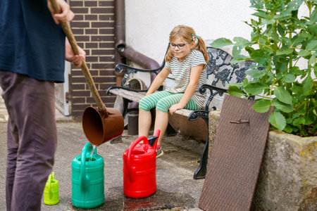 Little girl watering flowers in the garden. child waiting for father filling water in cans. Save water, drought,heat concept. Environment in europe or world. Rain waterの写真素材