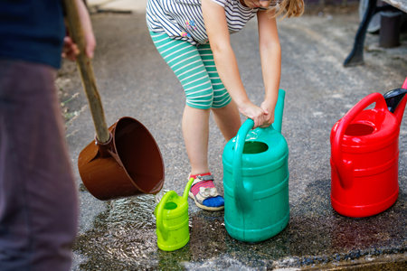 Little girl watering flowers in the garden. child waiting for father filling water in cans. Save water, drought,heat concept. Environment in europe or world. Rain waterの写真素材
