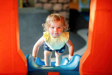 Happy blond little toddler girl having fun and sliding on indoor playground at daycare or nursery. Positive funny baby child smiling. Healthy girl climbing on slide.の写真素材