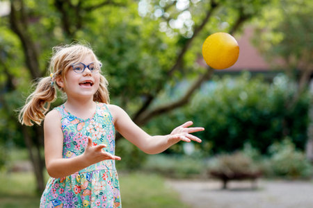 Little preschool girl with eyeglasses playing with ball outdoors. Happy smiling child catching and throwing, laughing and making sports. Active leisure with children and kids. Summer day on backyardの写真素材