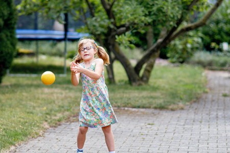 Little preschool girl with eyeglasses playing with ball outdoors. Happy smiling child catching and throwing, laughing and making sports. Active leisure with children and kids. Summer day on backyardの写真素材