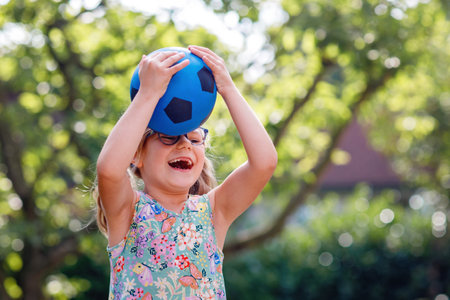 Little preschool girl with eyeglasses playing with ball outdoors. Happy smiling child catching and throwing, laughing and making sports. Active leisure with children and kids. Summer day on backyardの写真素材