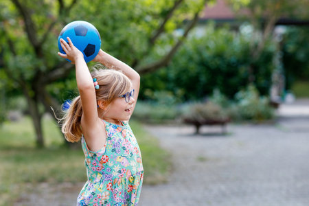 Little preschool girl with eyeglasses playing with ball outdoors. Happy smiling child catching and throwing, laughing and making sports. Active leisure with children and kids. Summer day on backyardの写真素材