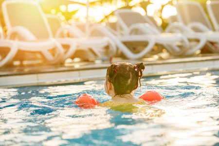 Little preschool girl with protective swimmies playing in outdoor swimming pool by sunset. Child learning to swim in outdoor pool, splashing with water, laughing and having fun. Family vacations.の写真素材