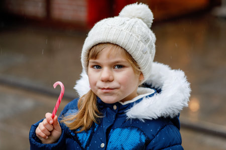 Little cute preschool girl with candy cane from a sweets stand on Christmas market. Happy child on traditional family market in Germany. Preschooler in colorful winter clothes during snowfallの写真素材