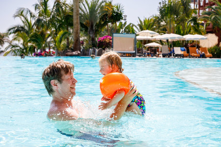 Cute happy little toddler girl and father in the pool and having fun on family vacations in a hotel resort. Healthy child and man playing in water. Baby daughter in colorful fashion swimsuit.の写真素材