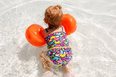 Little toddler girl with protective swimmies playing in outdoor swimming pool by sunset. Baby Child learning to swim in outdoor pool, splashing with water, laughing and having fun. Family vacations.の写真素材