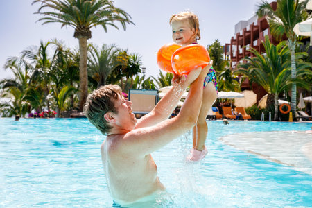Cute happy little toddler girl and father in the pool and having fun on family vacations in a hotel resort. Healthy child and man playing in water. Baby daughter in colorful fashion swimsuitの写真素材
