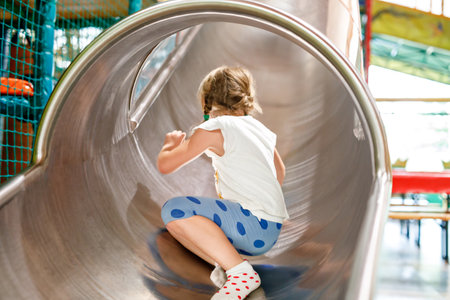 Little preschool girl riding from childrens slides on playground. Happy smiling child having fun in game center for kids.の写真素材