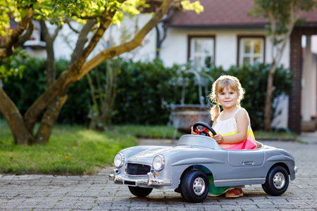 Little adorable toddler girl driving big vintage toy car and having fun with playing outdoors. Gorgeous happy healthy child enjoying warm summer day. Smiling stunning kid playing in domestic gardenの写真素材