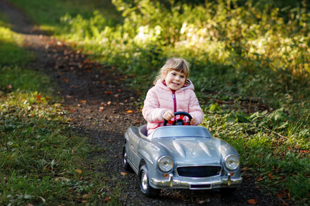 Little preschool girl driving big vintage toy car. Happy child having fun with playing outdoors. Active preschooler child enjoying warm autumn day in forest. Smiling stunning kid playingの写真素材