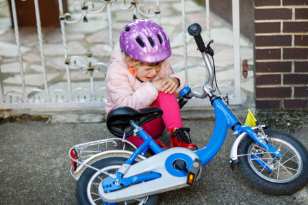 Cute little girl sitting on the ground after falling off her bike. Upset crying preschool child with safe helmet getting hurt while riding a bicycle. Active family leisure with kids.の写真素材