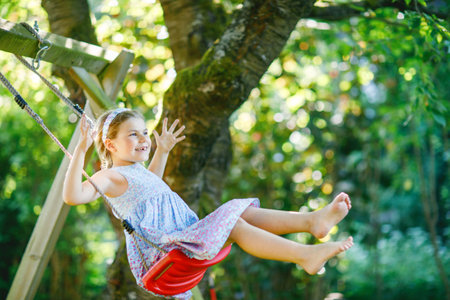 Happy little preschool girl having fun on swing in domestic garden. Cute healthy child swinging under blooming trees on sunny spring day. Kid laughing and crying.の写真素材