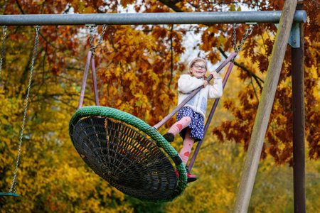 Happy child girl on swing in fall. Little kid playing in the autumn on playground, swinging and having fun. Autumnal trees on background. School backyard.の写真素材
