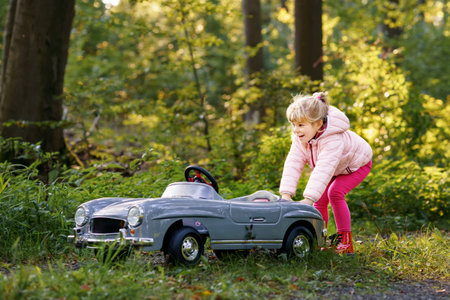 Little preschool girl driving big vintage toy car. Happy child having fun with playing outdoors. Active preschooler child enjoying warm autumn day in forest. Smiling stunning kid playingの写真素材