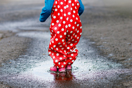 Little toddler girl wearing rain boots and trousers and walking during sleet, rain on cold day. Baby child having fun with jumping in a puddle. Active outdoors leisure and activity with little kids.の写真素材