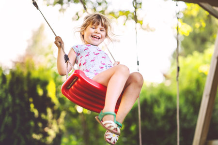 Happy little toddler girl having fun on swing in domestic garden. Smiling positive healthy child...