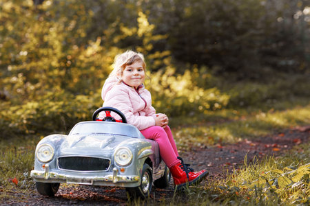 Little preschool girl driving big vintage toy car. Happy child having fun with playing outdoors. Active preschooler child enjoying warm autumn day in forest. Smiling stunning kid playingの写真素材