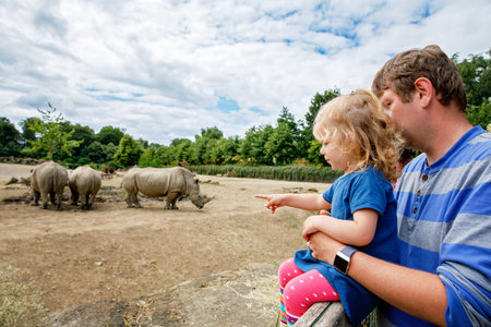 Cute adorable toddler girl and father watching wild rhinos in zoo. Happy baby child, daughter and dad, family having fun together with animals safari park on warm summer day. Irelandの写真素材