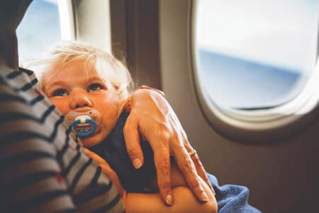 Woman traveling with little child by airplane. Sad tired toddler girl sitting with mum by aircraft window. Motherhood concept. Crying baby.の写真素材