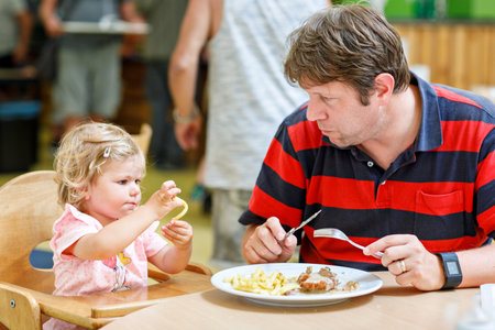 Toddler girl eating healthy vegetables and unhealthy french fries potatoes. Cute happy baby child taking food from parents dish in restaurant. Father eating in fast food restaurant with daughterの写真素材