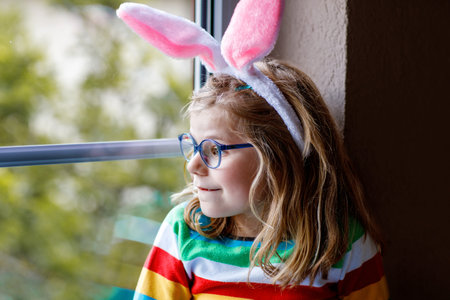 Portrait of a happy little girl with bunny ears looking outside sitting by a window. Easter holiday....