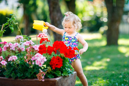 Cute little baby girl in colorful swimsuit watering plants and blossoming flowers in domestic garden on hot summer day. Adorable toddler child having fun with playing with water and caの写真素材