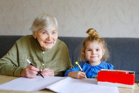 Beautiful toddler girl and grand grandmother drawing together pictures with felt pens at home. Cute child and senior woman having fun together. Happy family indoorsの写真素材
