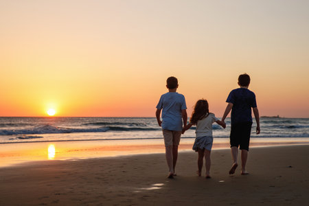 Three kids silhouettes running and jumping on beach at sunset. happy family, two school boys and one little preschool girl. Siblings having fun together. Bonding and family vacation.の写真素材