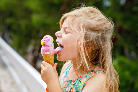 Happy preschool girl eating colorful ice cream in waffle cone on sunny summer day. Little toddler...