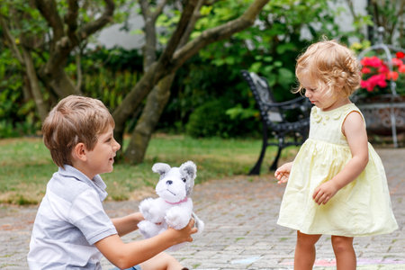 Little boy playing with toddler sister outdoors. Sibling drawing with chalks and play with soft toy. Happy girl and kid boy in summer.の写真素材
