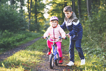Cute little preschool girl in safety helmet riding bicycle. School kid boy, brother teaching happy healthy sister child cycling and having fun with learning bike. Active siblings family outdoors.の写真素材