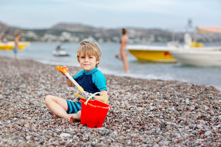 Adorable little blond kid boy building pebble stone castle on beach. Funny child playing with bucket and shovel. Vacations, summer, travel concept. Toddler enjoying summer vacations on sea.の写真素材