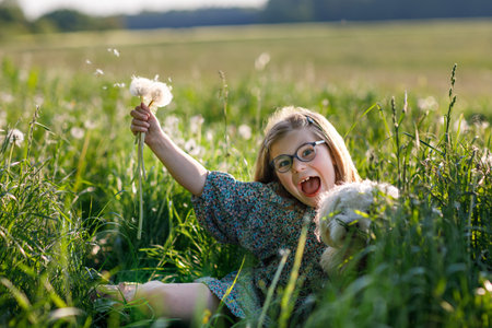 Adorable cute little girl blowing on a dandelion flower on the nature in the summer. Happy school child with eyeglasses holding Maltese dog, having fun with playing. Bright sunset light, active kid.の写真素材