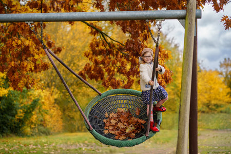 Happy child girl on swing in fall. Little kid playing in the autumn on playground, swinging and having fun. Autumnal trees on background. School backyard.の写真素材